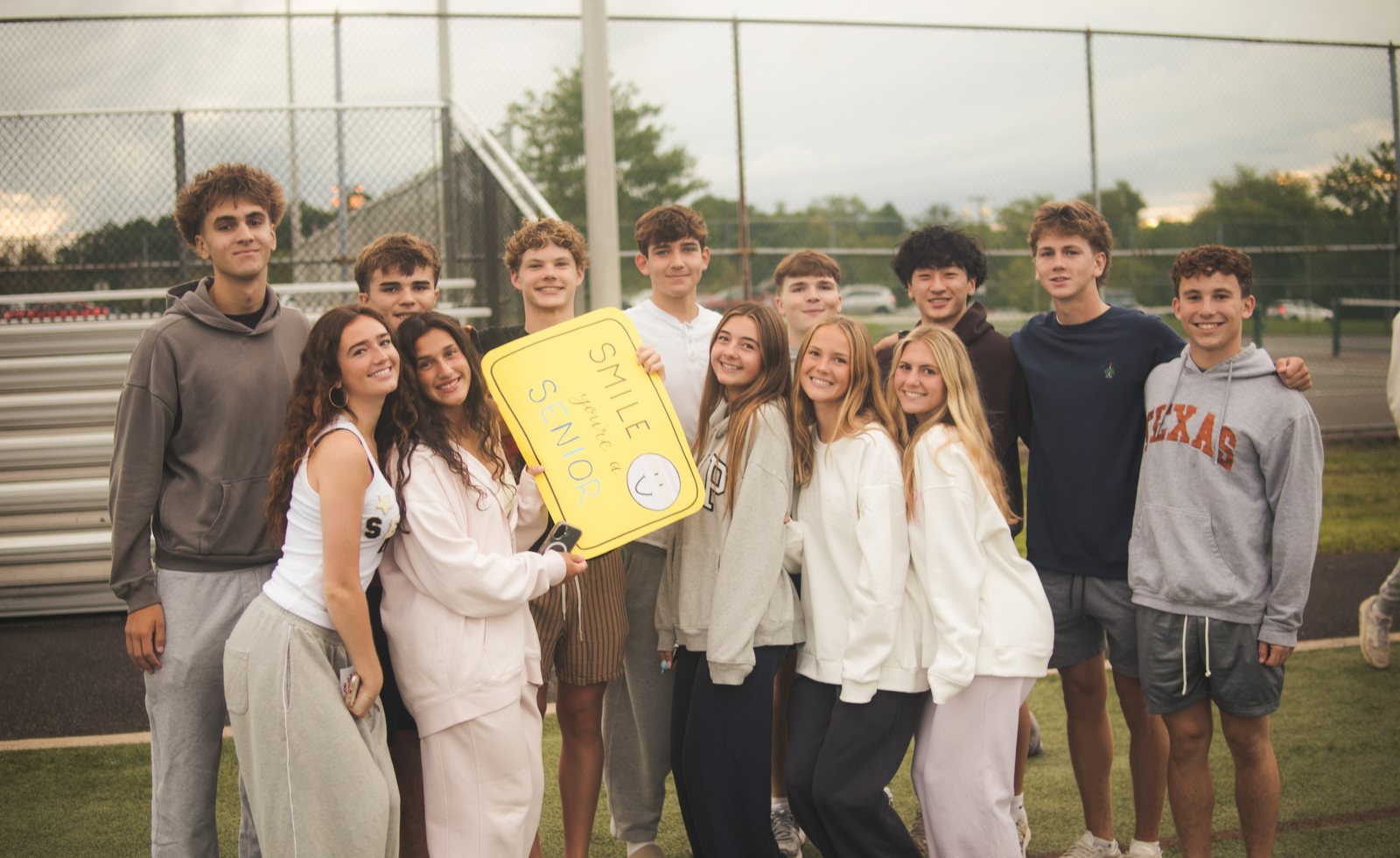 Seniors posing on the turf field by the fence, two holding a yellow sign that reads Smile, you’re a senior