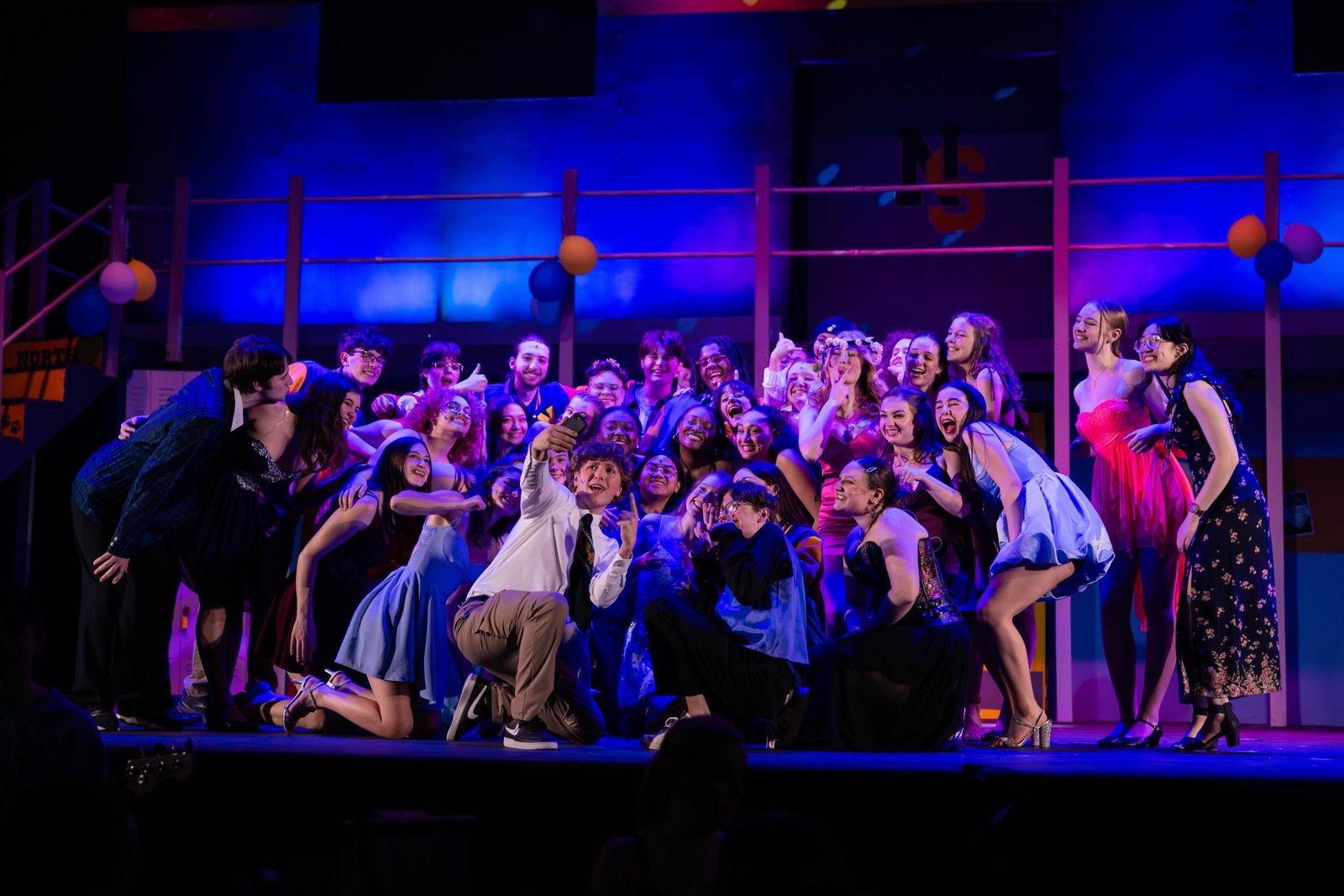 A large cast in costume crowded together on a lit stage set, smiling