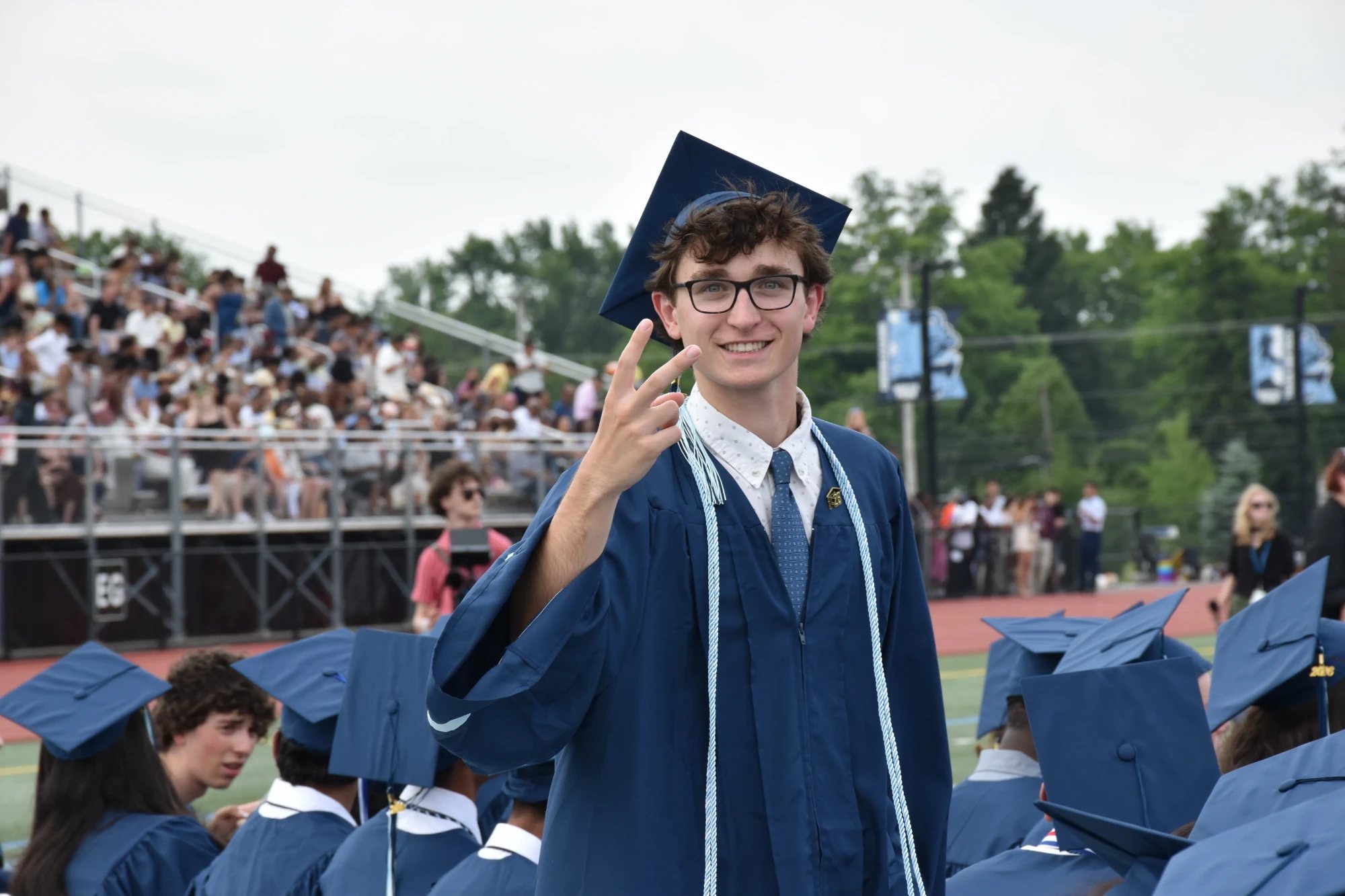 A graduate in a navy cap and gown smiling on the stadium field, the stands full behind