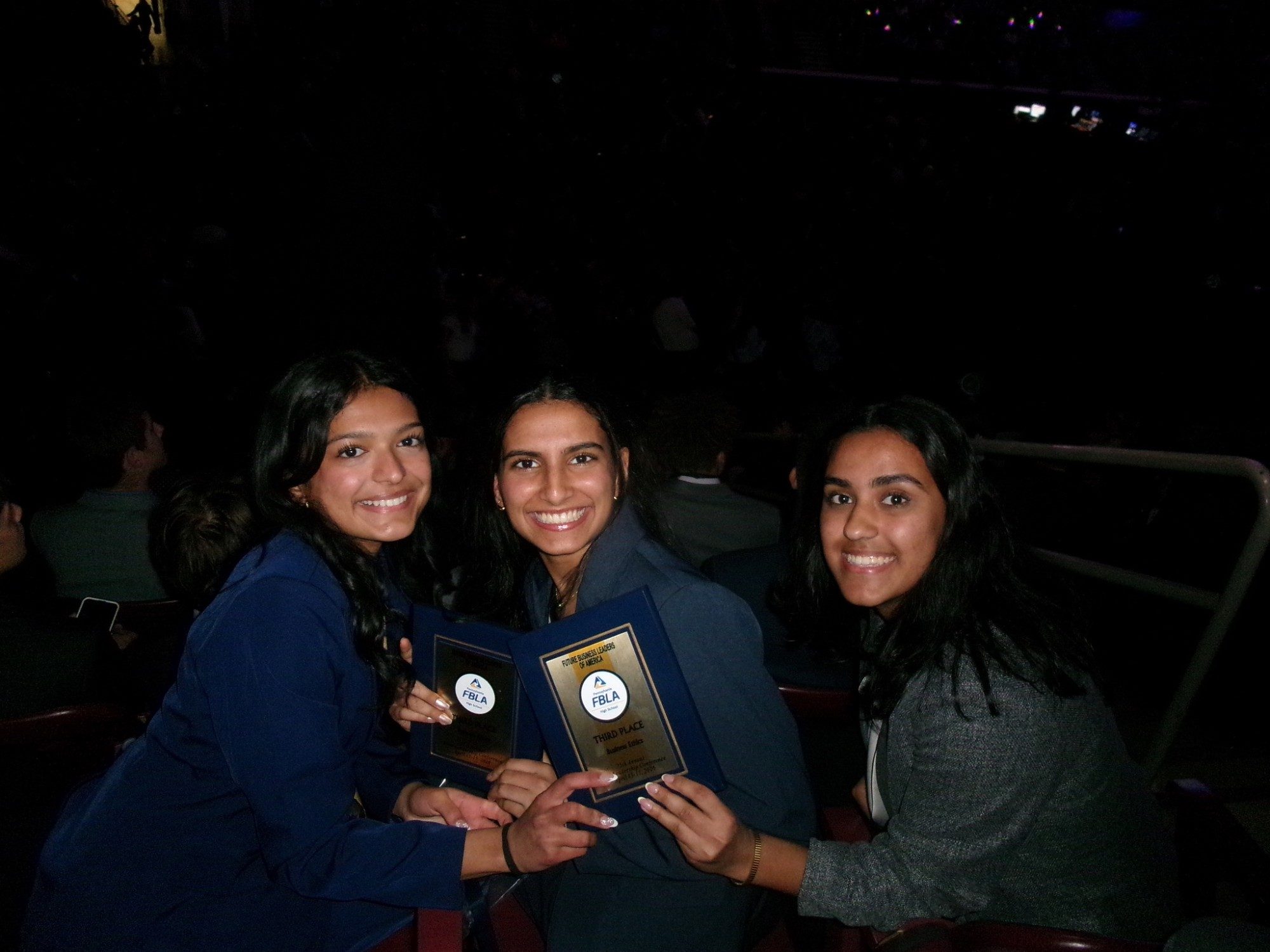 Three members in the arena seats, leaning together and holding up their award plaques