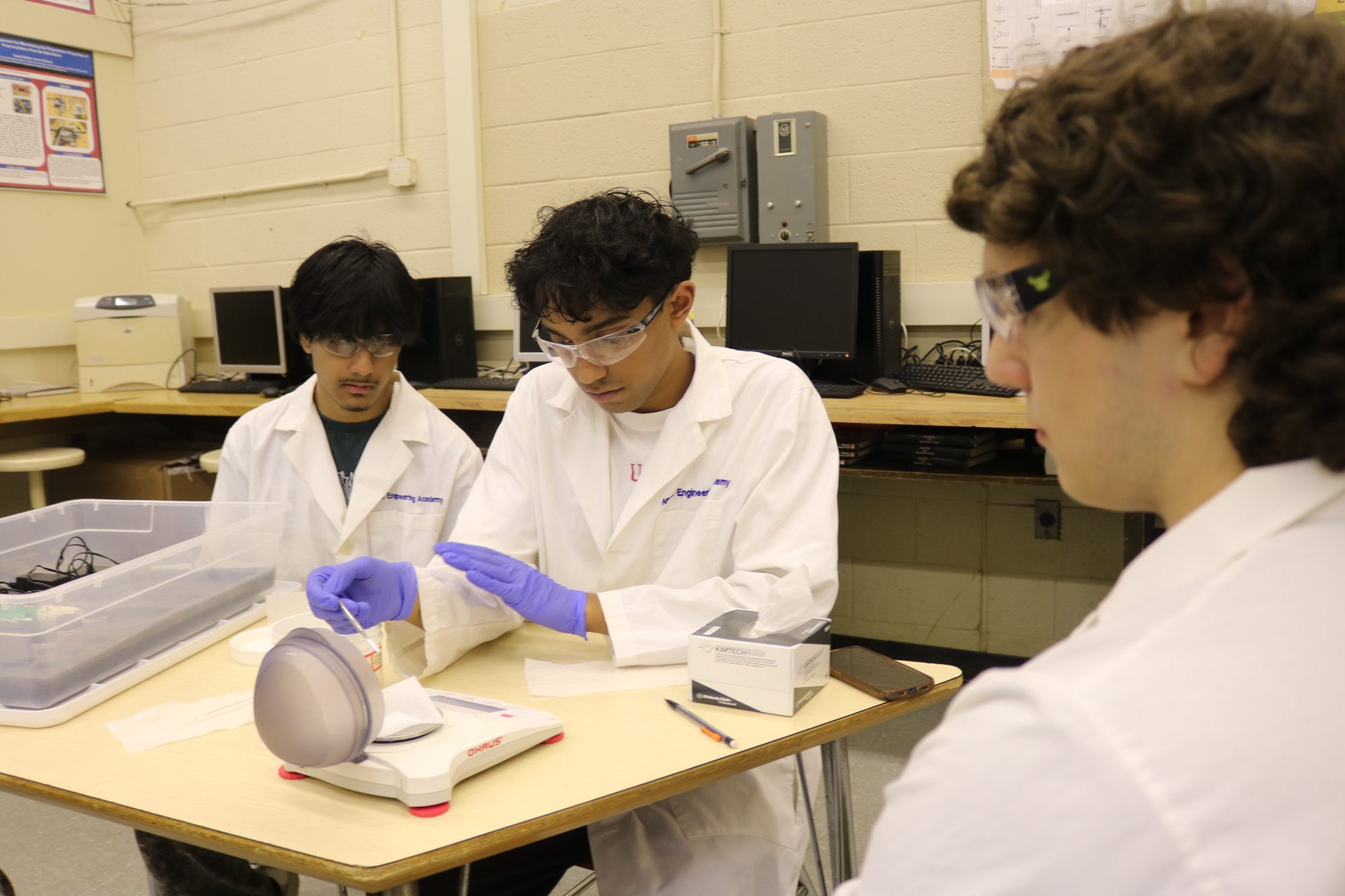 Three Engineering Academy students in lab coats and safety glasses weighing material at a lab bench