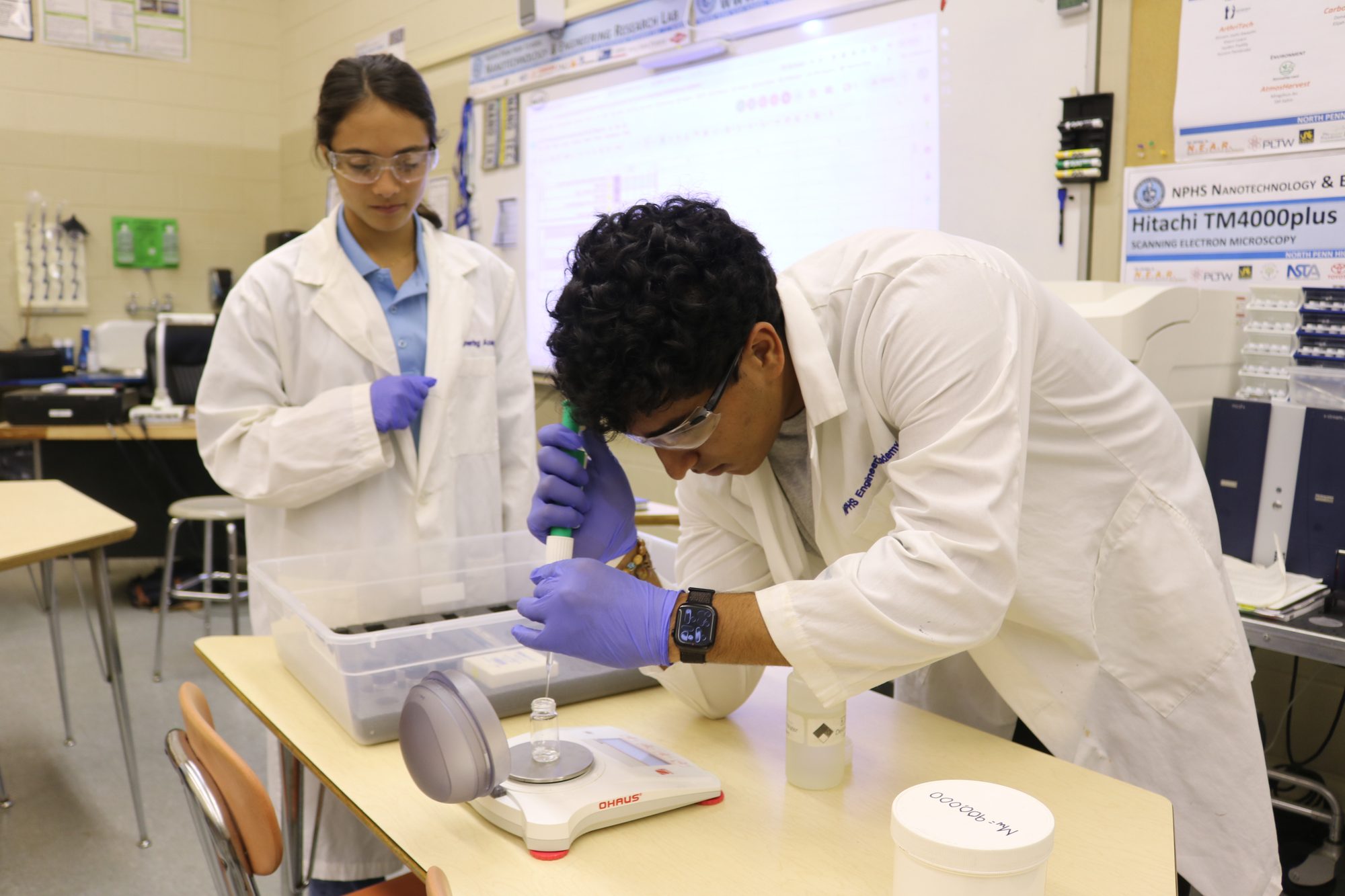 Two students in lab coats and safety glasses: one pipettes liquid into a small vial on a balance while the other watches